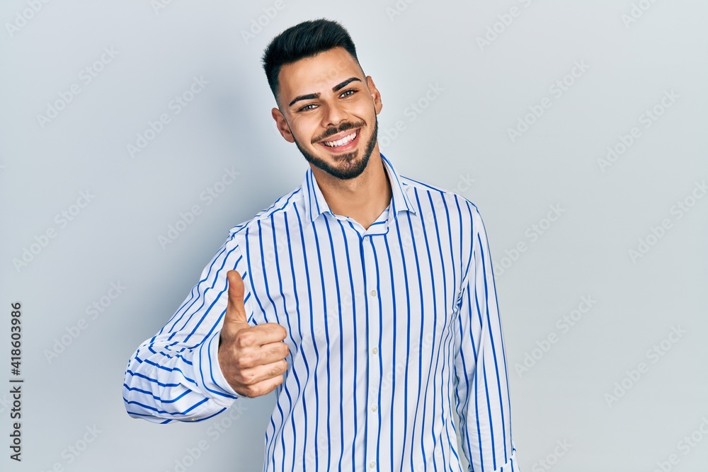 Fototapeta premium Young hispanic man with beard wearing casual striped shirt doing happy thumbs up gesture with hand. approving expression looking at the camera showing success.