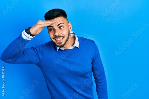 Young hispanic man with beard wearing casual blue sweater very happy and smiling looking far away with hand over head. searching concept.