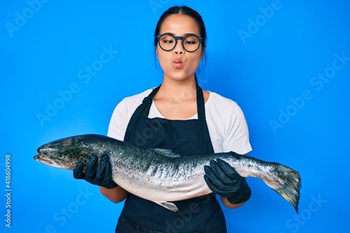 Young beautiful asian girl fishmonger selling fresh raw salmon making fish face with mouth and squinting eyes, crazy and comical.