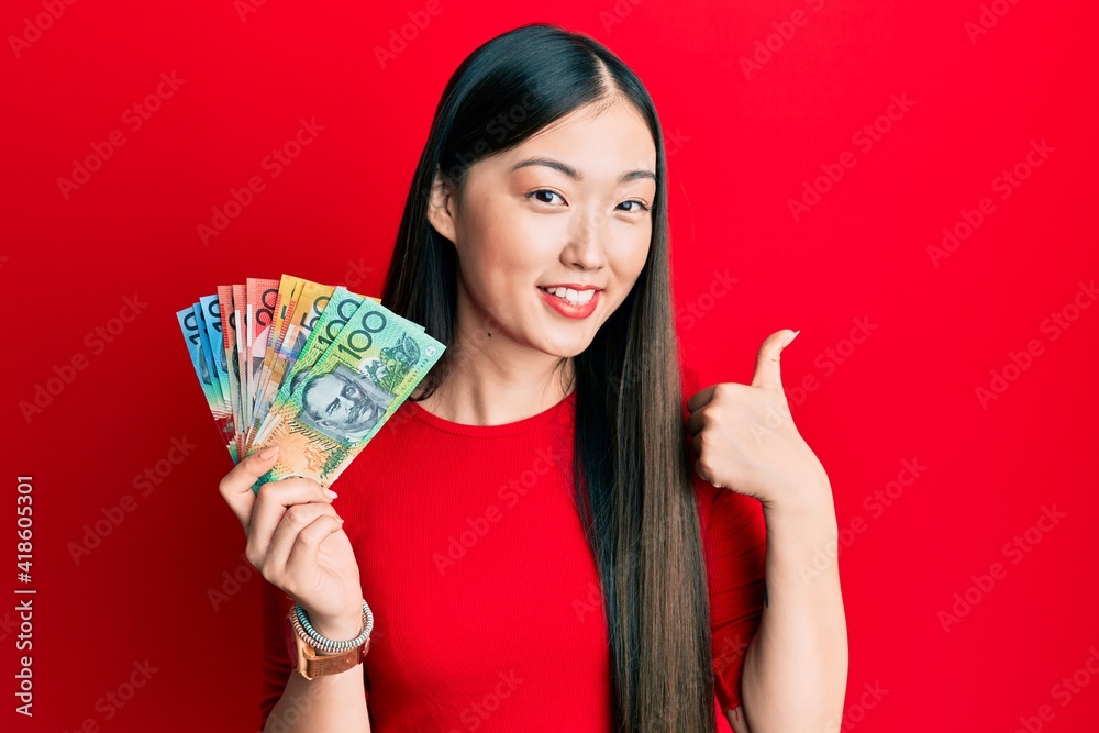 Young chinese woman holding australian dollars smiling happy and positive, thumb up doing excellent and approval sign Young chinese woman holding australian dollars smiling happy and positive, thumb up doing excellent and approval sign
