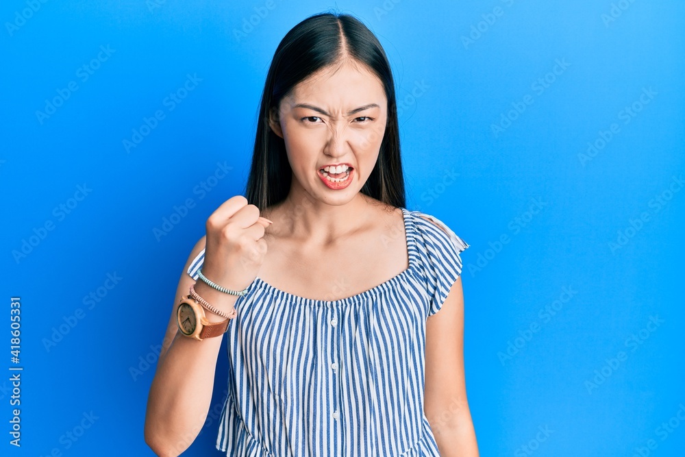 Young chinese woman wearing casual striped t-shirt angry and mad raising fist frustrated and furious while shouting with anger. rage and aggressive concept. Young chinese woman wearing casual striped t-shirt angry and mad raising fist frustrated and furious while shouting with anger. rage and aggressive concept.