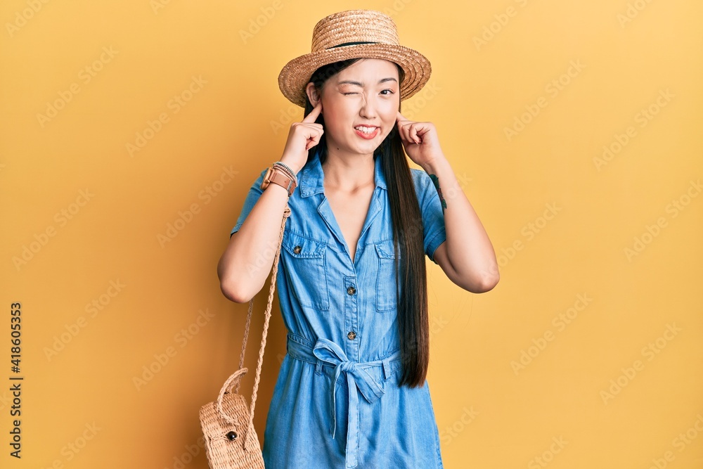 Young chinese woman wearing summer hat covering ears with fingers with annoyed expression for the noise of loud music. deaf concept.