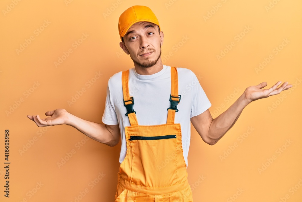 Hispanic young man wearing handyman uniform clueless and confused expression with arms and hands raised. doubt concept. Hispanic young man wearing handyman uniform clueless and confused expression with arms and hands raised. doubt concept.
