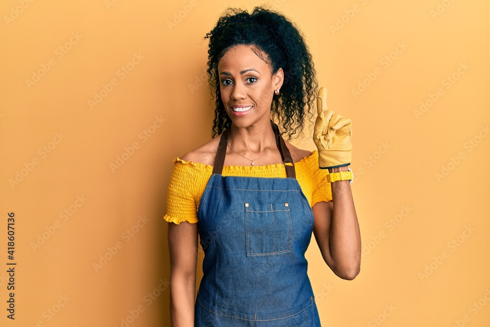Middle age african american woman wearing professional apron showing and pointing up with finger number one while smiling confident and happy. Middle age african american woman wearing professional apron showing and pointing up with finger number one while smiling confident and happy.