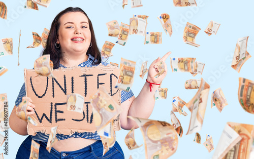 Young plus size woman holding united we stand banner smiling happy pointing with hand and finger to the side