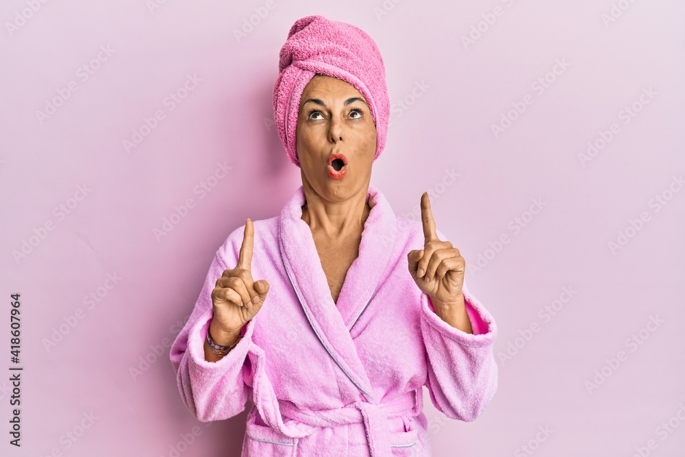 Middle age hispanic woman wearing shower towel cap and bathrobe amazed and surprised looking up and pointing with fingers and raised arms. Middle age hispanic woman wearing shower towel cap and bathrobe amazed and surprised looking up and pointing with fingers and raised arms.