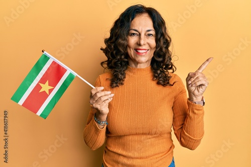 Middle age hispanic woman holding suriname flag smiling happy pointing with hand and finger to the side