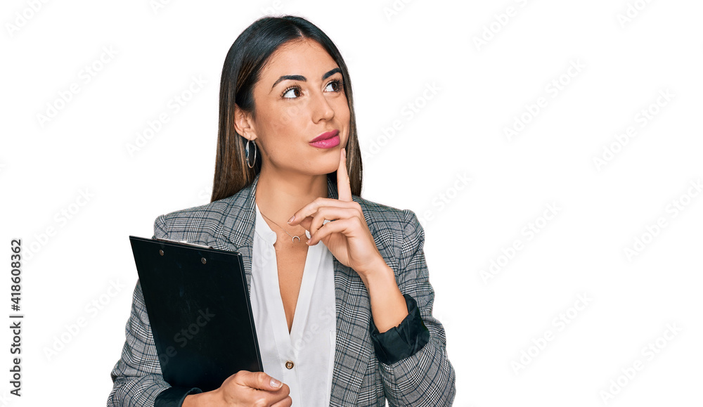 Young hispanic woman wearing business clothes holding clipboard serious face thinking about question with hand on chin, thoughtful about confusing idea Young hispanic woman wearing business clothes holding clipboard serious face thinking about question with hand on chin, thoughtful about confusing idea