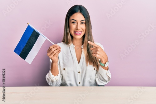 Young hispanic woman holding estonia flag sitting on the table smiling happy pointing with hand and finger