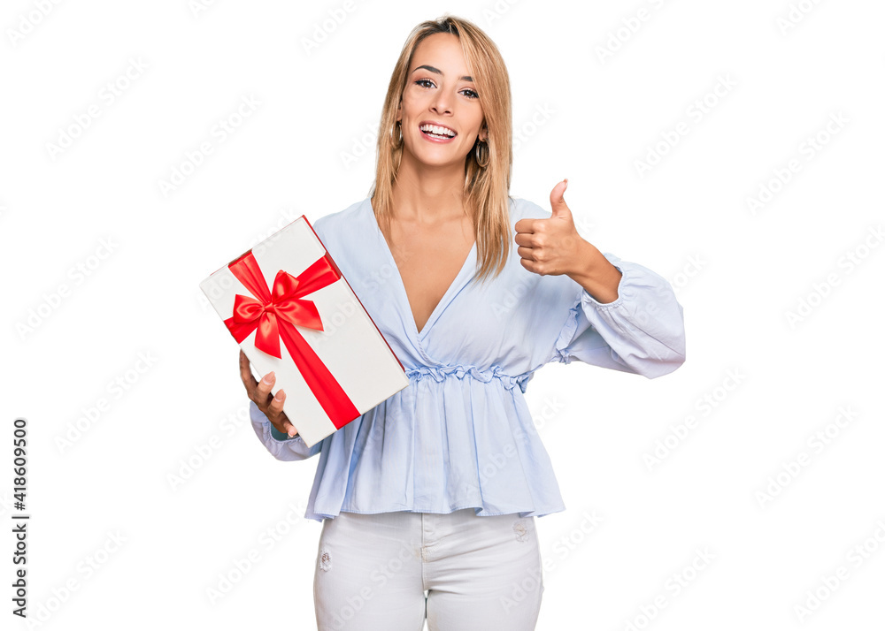 Beautiful blonde woman holding gift smiling happy and positive, thumb up doing excellent and approval sign Beautiful blonde woman holding gift smiling happy and positive, thumb up doing excellent and approval sign