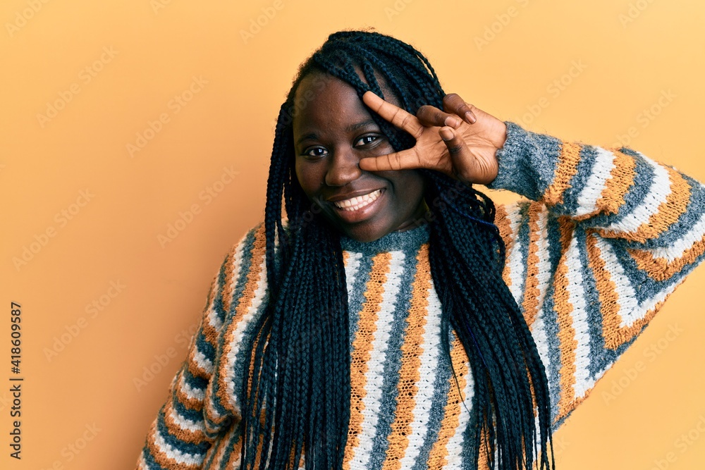 Young black woman with braids wearing casual winter sweater doing peace symbol with fingers over face, smiling cheerful showing victory Young black woman with braids wearing casual winter sweater doing peace symbol with fingers over face, smiling cheerful showing victory