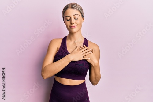 Beautiful blonde woman wearing sportswear over pink background smiling with hands on chest with closed eyes and grateful gesture on face. health concept.