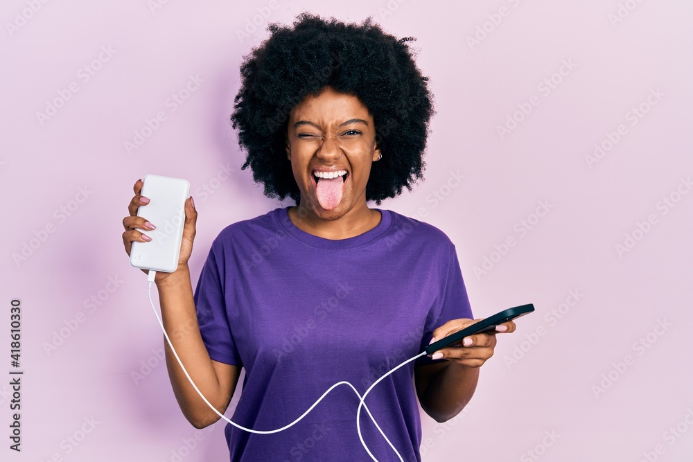 Young african american woman charging smartphone using portable battery sticking tongue out happy with funny expression. - obrazy, fototapety, plakaty 