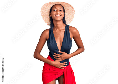 Young african american woman wearing swimsuit and summer hat smiling and laughing hard out loud because funny crazy joke with hands on body. photo