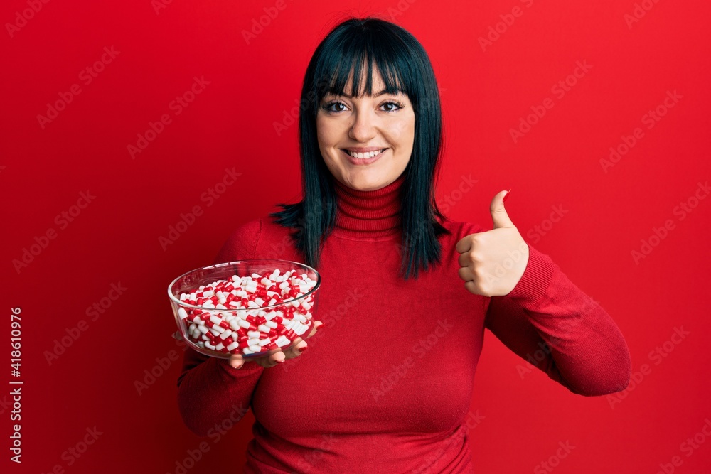 Young hispanic woman holding bowl of pills smiling happy and positive, thumb up doing excellent and approval sign Young hispanic woman holding bowl of pills smiling happy and positive, thumb up doing excellent and approval sign