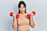 Young hispanic woman wearing sportswear using dumbbells making fish face with mouth and squinting eyes, crazy and comical.
