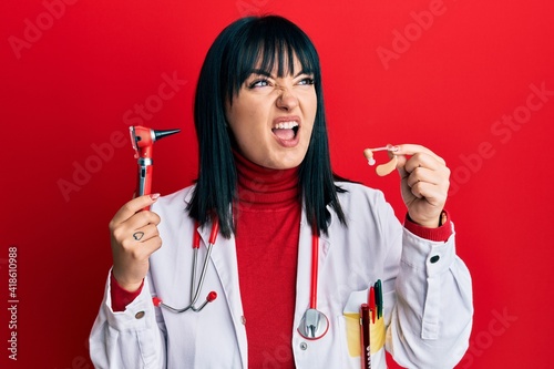 Young hispanic doctor woman holding deafness headset and otoscope angry and mad screaming frustrated and furious, shouting with anger. rage and aggressive concept.