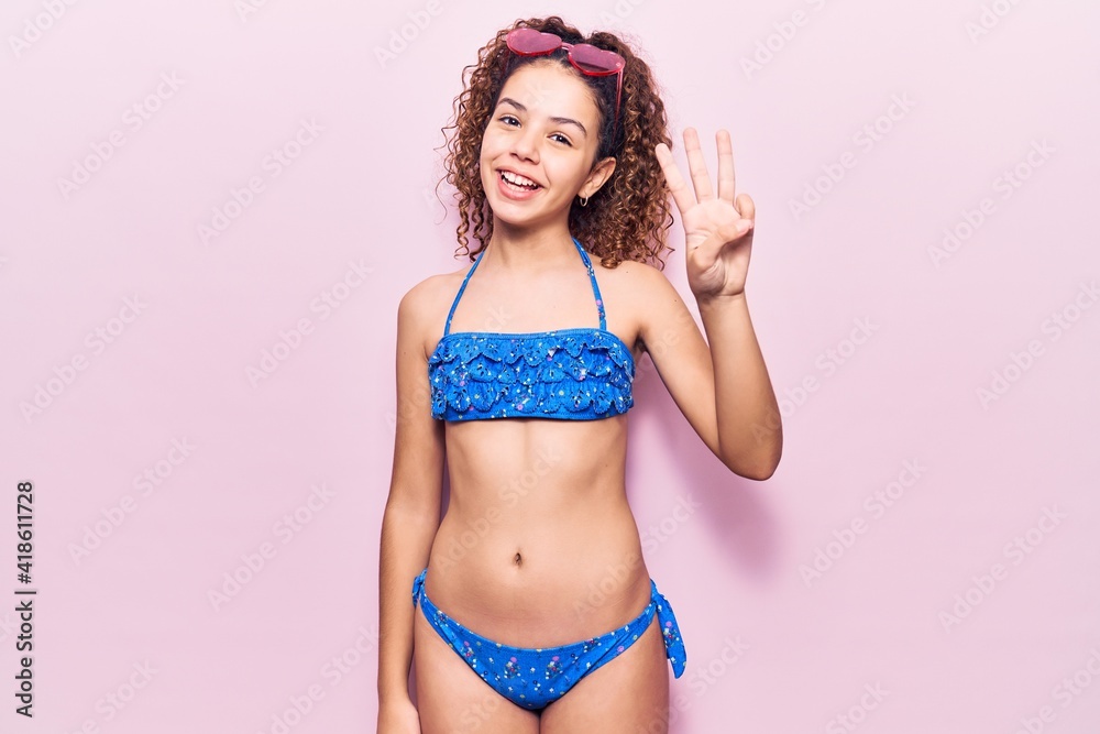Naklejka premium Beautiful kid girl with curly hair wearing bikini and sunglasses showing and pointing up with fingers number three while smiling confident and happy.