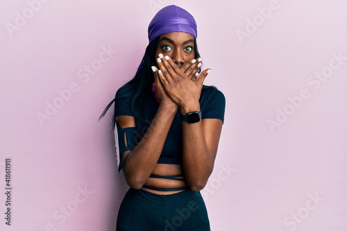 Young african american woman wearing gym clothes and using headphones shocked covering mouth with hands for mistake. secret concept.