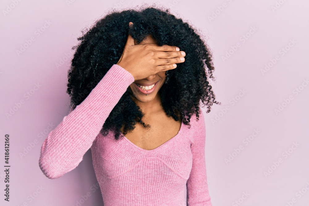 African american woman with afro hair wearing casual pink shirt smiling and laughing with hand on face covering eyes for surprise. blind concept. African american woman with afro hair wearing casual pink shirt smiling and laughing with hand on face covering eyes for surprise. blind concept.