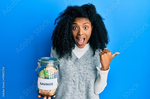 African american woman with afro hair holding savings jar with south african rands money pointing thumb up to the side smiling happy with open mouth photo