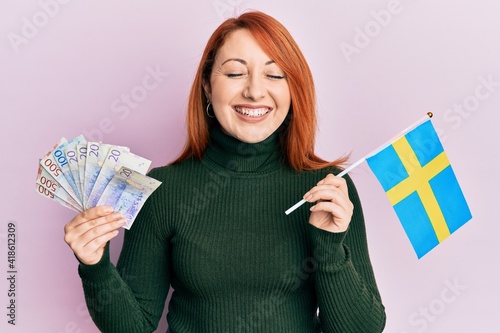 Beautiful redhead woman holding 20 swedish krona banknotes and sweden flag smiling and laughing hard out loud because funny crazy joke. photo