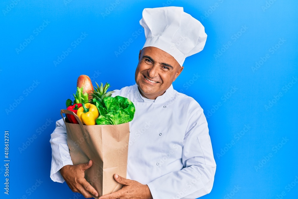 Mature middle east man professional chef holding bag of groceries looking positive and happy standing and smiling with a confident smile showing teeth Mature middle east man professional chef holding bag of groceries looking positive and happy standing and smiling with a confident smile showing teeth