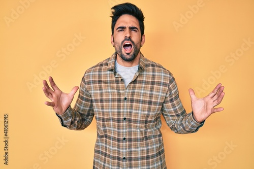 Handsome hispanic man with beard wearing casual clothes crazy and mad shouting and yelling with aggressive expression and arms raised. frustration concept.