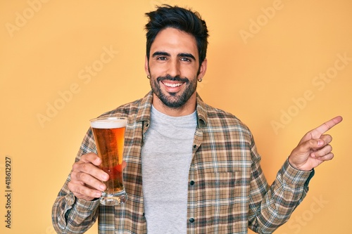 Handsome hispanic man with beard drinking a pint of beer smiling happy pointing with hand and finger to the side