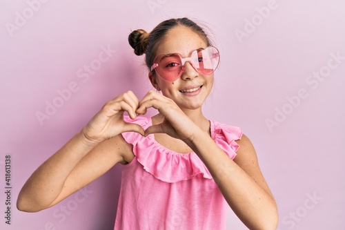 Beautiful brunette little girl wearing heart shaped sunglasses smiling in love doing heart symbol shape with hands. romantic concept.