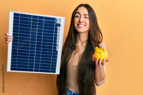 Foto Young hispanic girl holding photovoltaic solar panel and piggy bank winking looking at the camera with sexy expression, cheerful and happy face