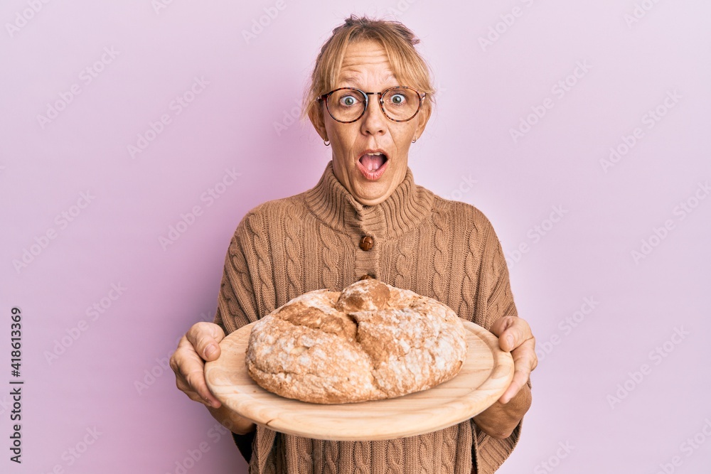 Middle age blonde woman holding bread afraid and shocked with surprise and amazed expression, fear and excited face. Middle age blonde woman holding bread afraid and shocked with surprise and amazed expression, fear and excited face.