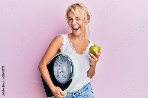 Young blonde girl holding weight machine and green apple winking looking at the camera with sexy expression  cheerful and happy face.