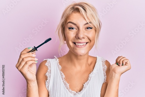 Young blonde girl holding eyelash curler screaming proud, celebrating victory and success very excited with raised arm