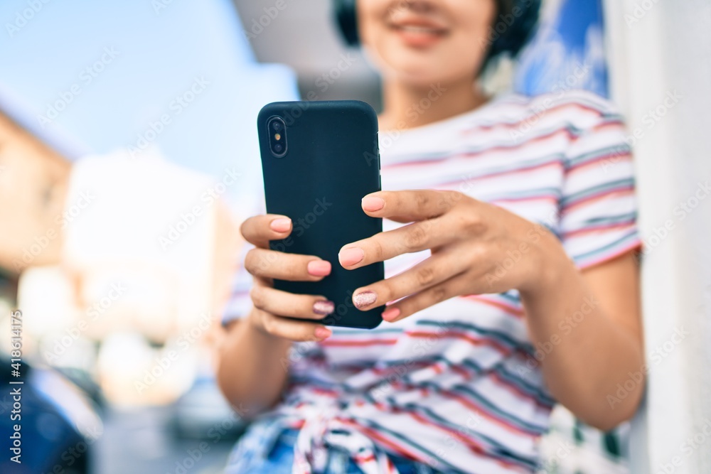 Fototapeta premium Young latin girl smiling happy using smartphone and headphones at the city