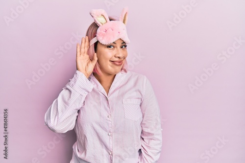 Hispanic woman with pink hair wearing sleep mask and pajama smiling with hand over ear listening an hearing to rumor or gossip. deafness concept.