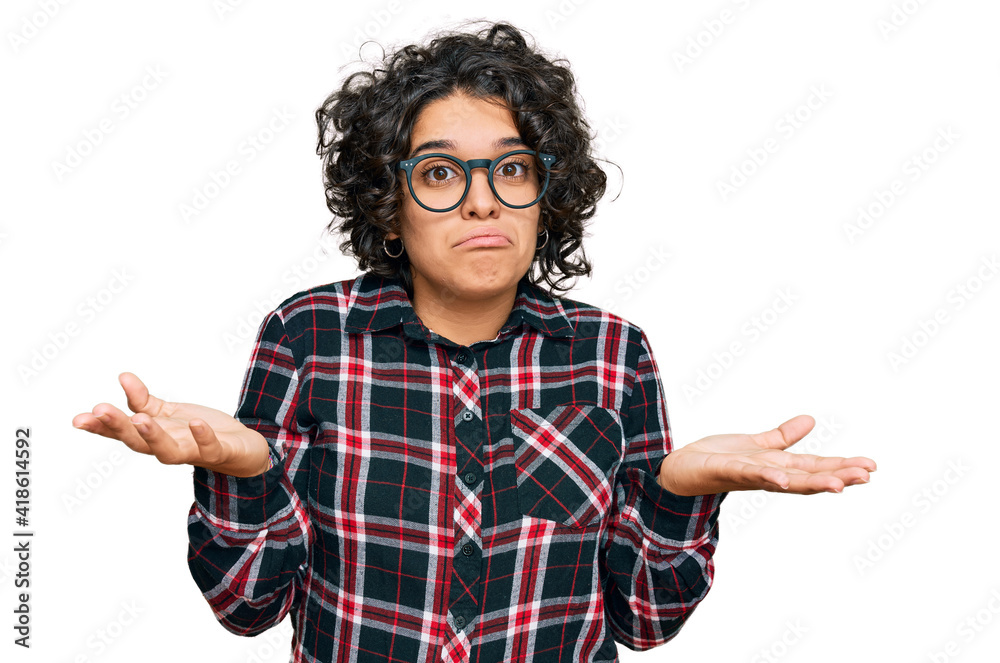 Young hispanic woman with curly hair wearing casual clothes and glasses clueless and confused expression with arms and hands raised. doubt concept. Young hispanic woman with curly hair wearing casual clothes and glasses clueless and confused expression with arms and hands raised. doubt concept.