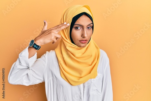 Young brunette arab woman wearing traditional islamic hijab scarf shooting and killing oneself pointing hand and fingers to head like gun, suicide gesture. © Krakenimages.com