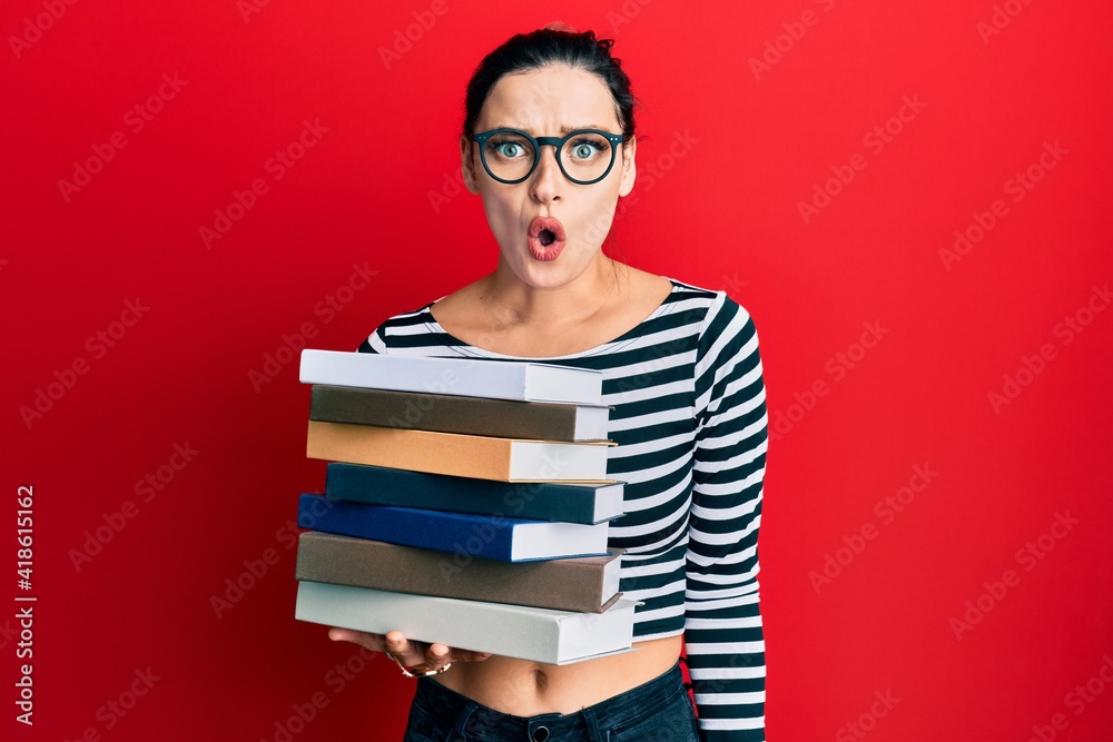 Young caucasian woman wearing glasses and holding books scared and amazed with open mouth for surprise, disbelief face Young caucasian woman wearing glasses and holding books scared and amazed with open mouth for surprise, disbelief face