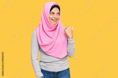 Young caucasian woman wearing traditional islamic hijab scarf smiling with happy face looking and pointing to the side with thumb up. © Krakenimages.com