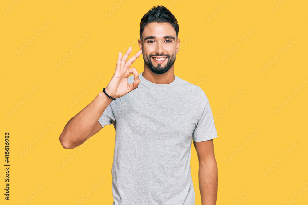 Young man with beard wearing casual grey tshirt smiling positive doing ok sign with hand and fingers. successful expression.