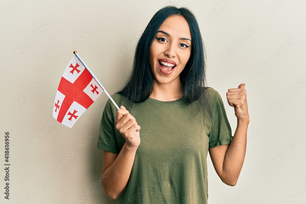 Young hispanic girl holding georgia flag pointing thumb up to the side smiling happy with open mouth Young hispanic girl holding georgia flag pointing thumb up to the side smiling happy with open mouth