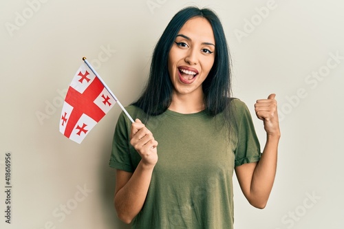 Young hispanic girl holding georgia flag pointing thumb up to the side smiling happy with open mouth