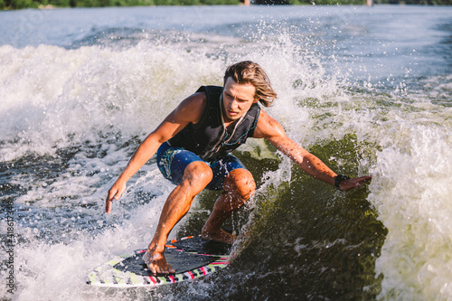 Beautiful long haired blond wakesurf in vest on board along waves of lake. Athletic male athlete wakes surfing in summer on river. Wakesurf water sport. Surfer on wave. Man surfing on the Surfboard