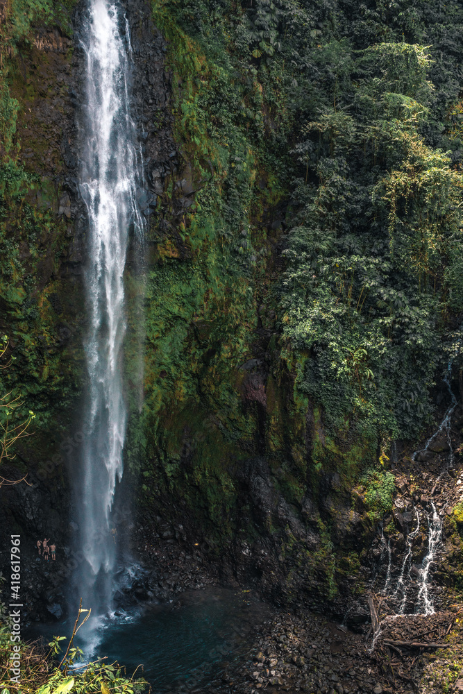 Huge waterfall with cold water falling from the green and leafy mountain with some tourists at the bottom Huge waterfall with cold water falling from the green and leafy mountain with some tourists at the bottom