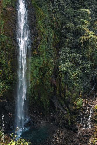 Huge waterfall with cold water falling from the green and leafy mountain with some tourists at the bottom