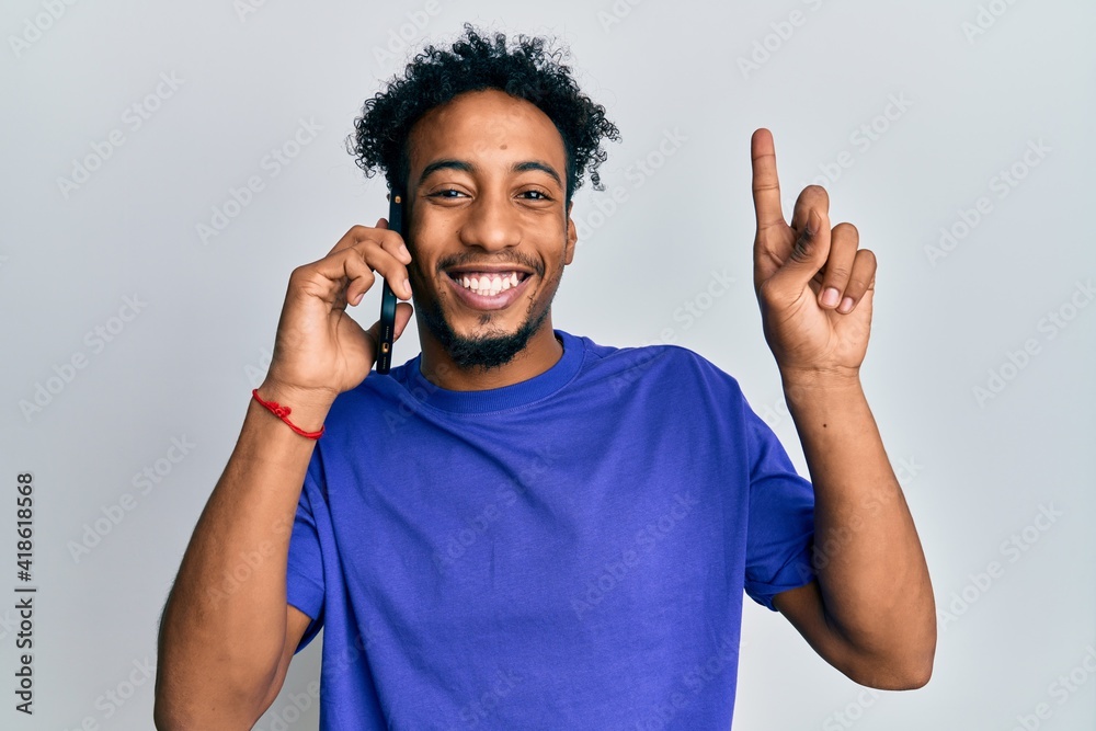 Fototapeta premium Young african american man with beard having conversation talking on the smartphone smiling with an idea or question pointing finger with happy face, number one