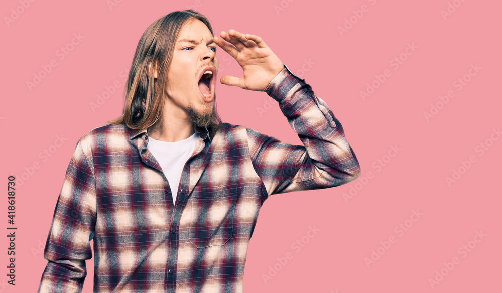 Handsome caucasian man with long hair wearing hipster shirt shouting and screaming loud to side with hand on mouth. communication concept. Handsome caucasian man with long hair wearing hipster shirt shouting and screaming loud to side with hand on mouth. communication concept.