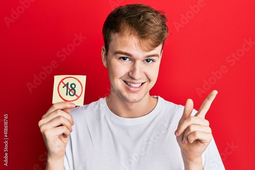 Young caucasian man holding under 18 prohibition sticker smiling happy pointing with hand and finger to the side photo