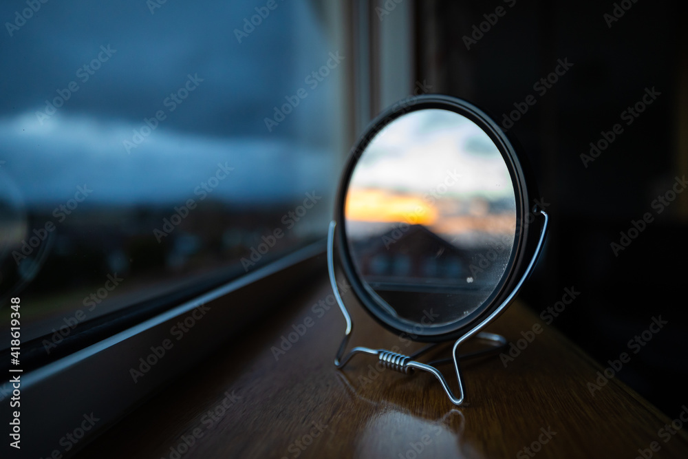 Small black makeup mirror sitting by a window reflecting the distant sunset. Blurred orange mixed in with strong blues. Creative sunset shot. Small black makeup mirror sitting by a window reflecting the distant sunset. Blurred orange mixed in with strong blues. Creative sunset shot.